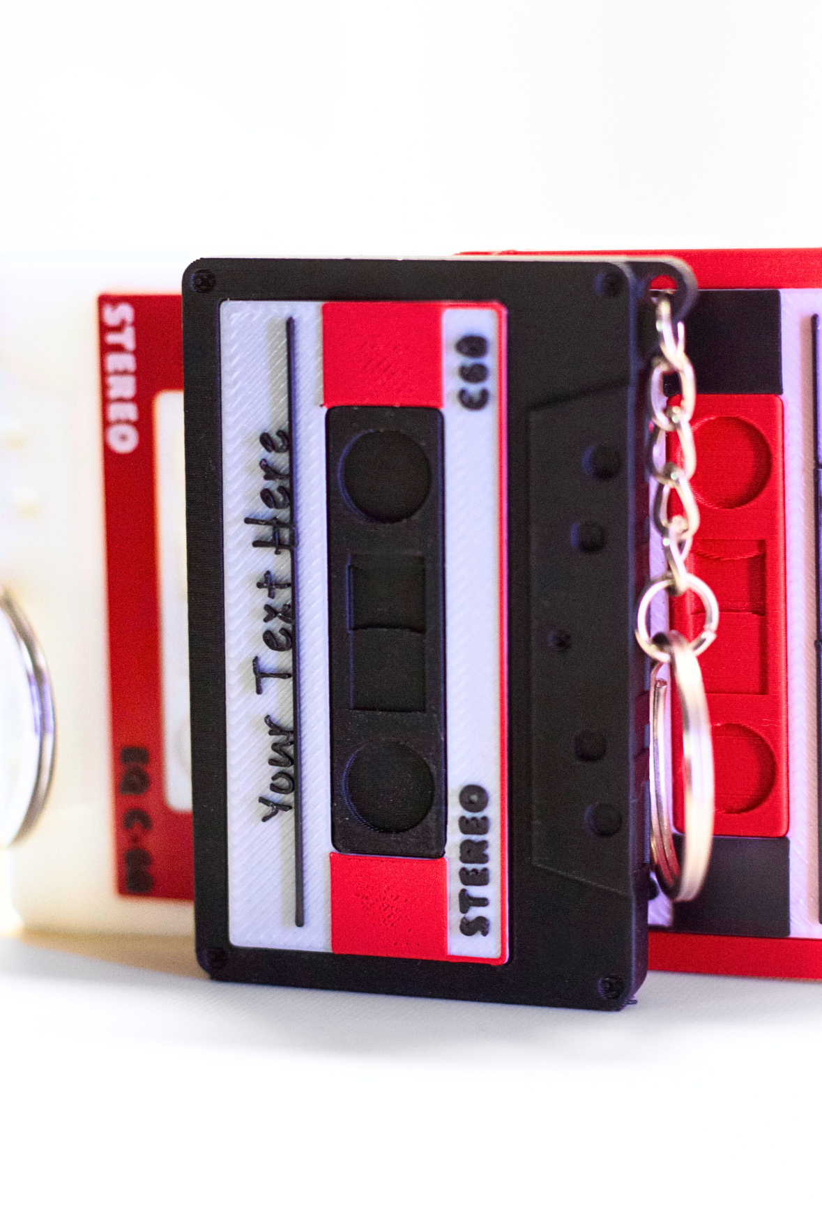 An image of a black cassette keychain with a red and white cassette keychain blurred in the background, against a white background. The label on the black cassette in the front of the image is white and red with black text. 